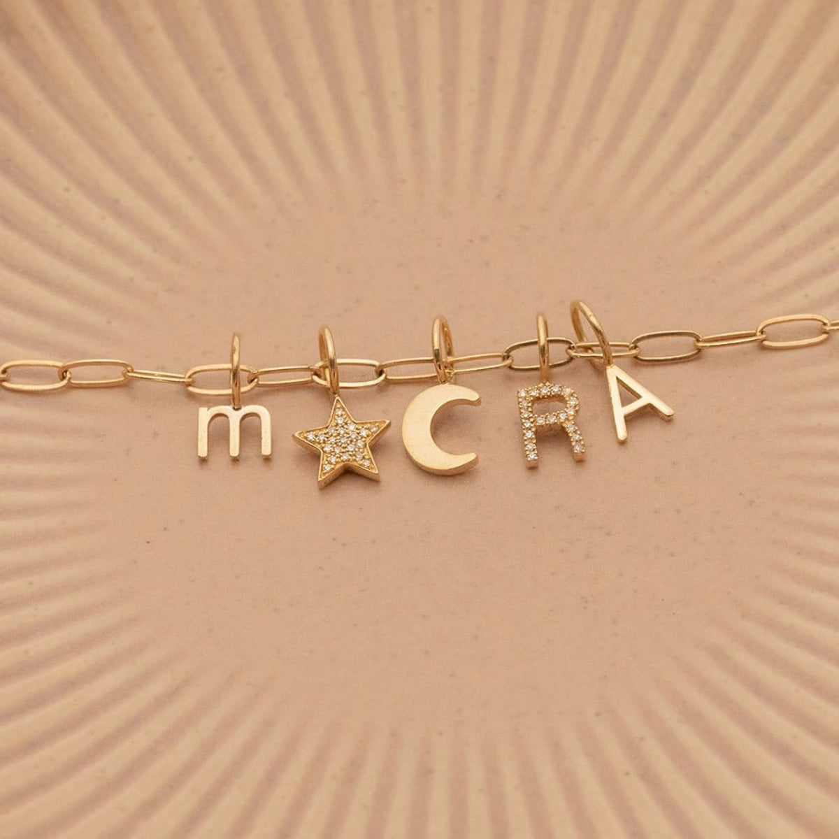 An Element Long Link Chain with a Petite Letter Charm in M, Charm Pendants in Moon and Pavé Star, Pavé Letter Charm in R and Gold Letter Charm in S against a beige background.