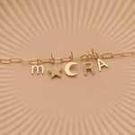 An Element Long Link Chain with a Petite Letter Charm in M, Charm Pendants in Moon and Pavé Star, Pavé Letter Charm in R and Gold Letter Charm in S against a beige background.