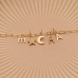 An Element Long Link Chain with a Petite Letter Charm in M, Charm Pendants in Moon and Pavé Star, Pavé Letter Charm in R and Gold Letter Charm in S against a beige background.