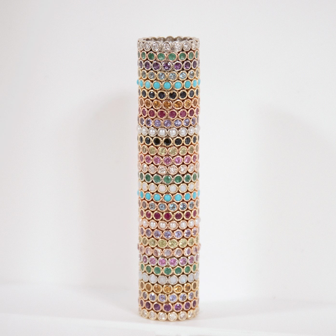 A gold-toned cylindrical colourful eternity rings in pink, green, blue, purple, and white, set against a plain white background.