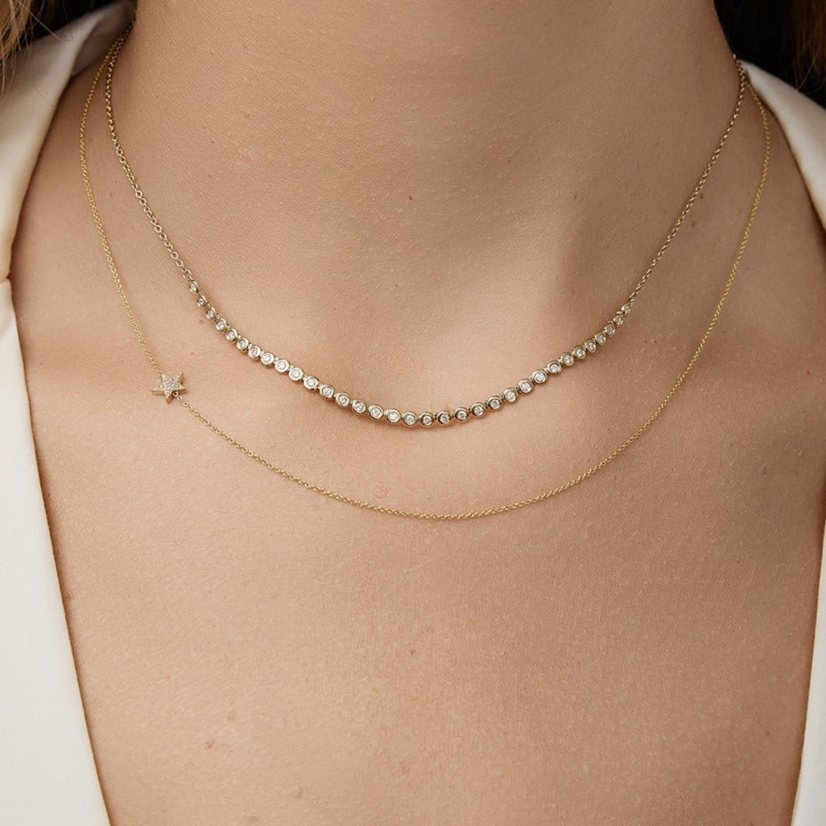 Close-up of a person's neck wearing The Everyday Diamond Necklace layered with a 14k Gold Asymmetrical Charm Necklace - Pavé Star in yellow gold, styled with a cream top. 