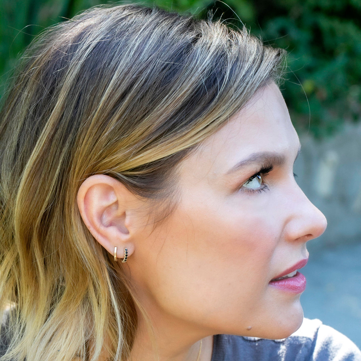Close-up of a woman's profile with blonde hair and earrings, blurred greenery in the background