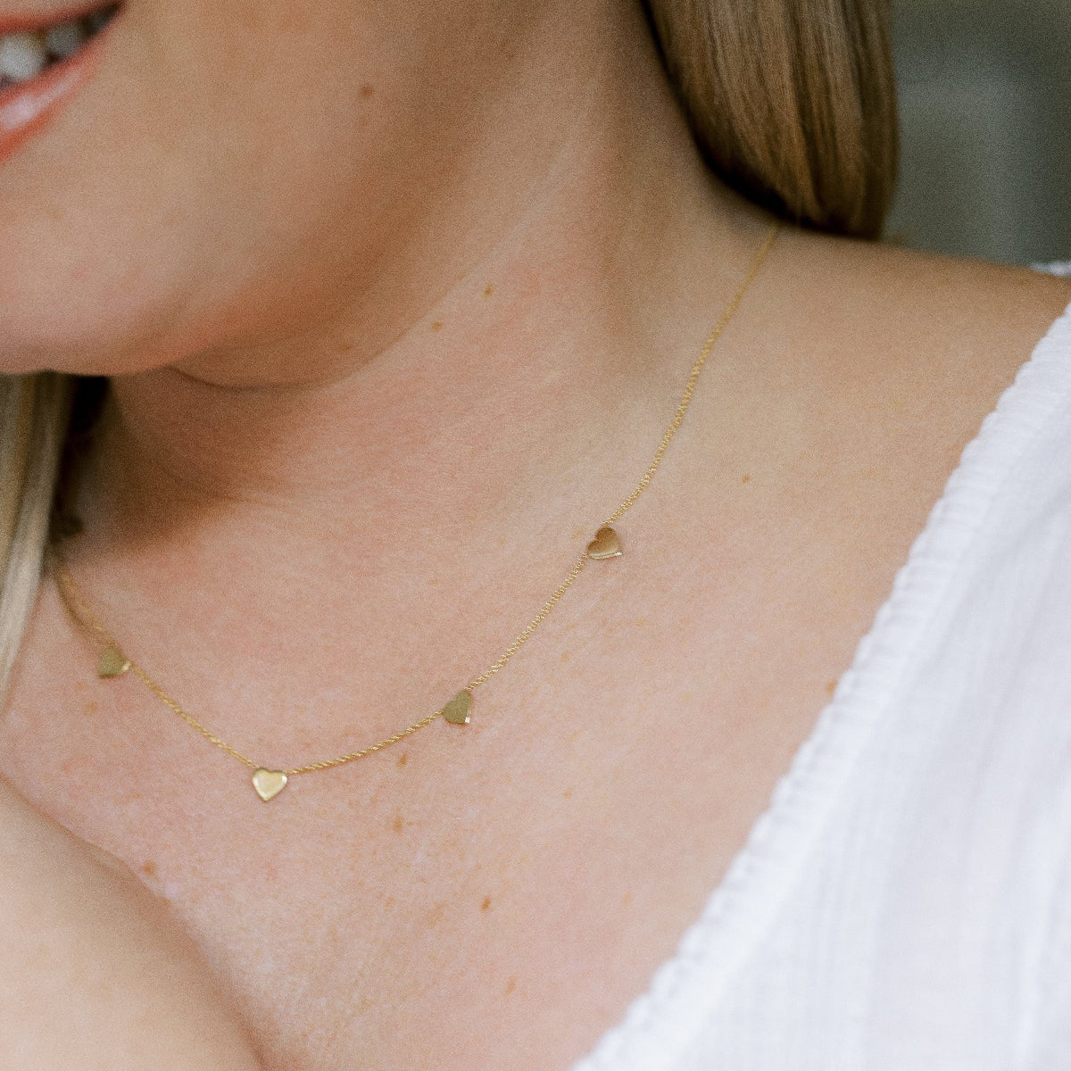 Close up of a  woman's neck wearing a 5 of Hearts Necklace in yellow gold with a white top and her blonde hair partially visible. 