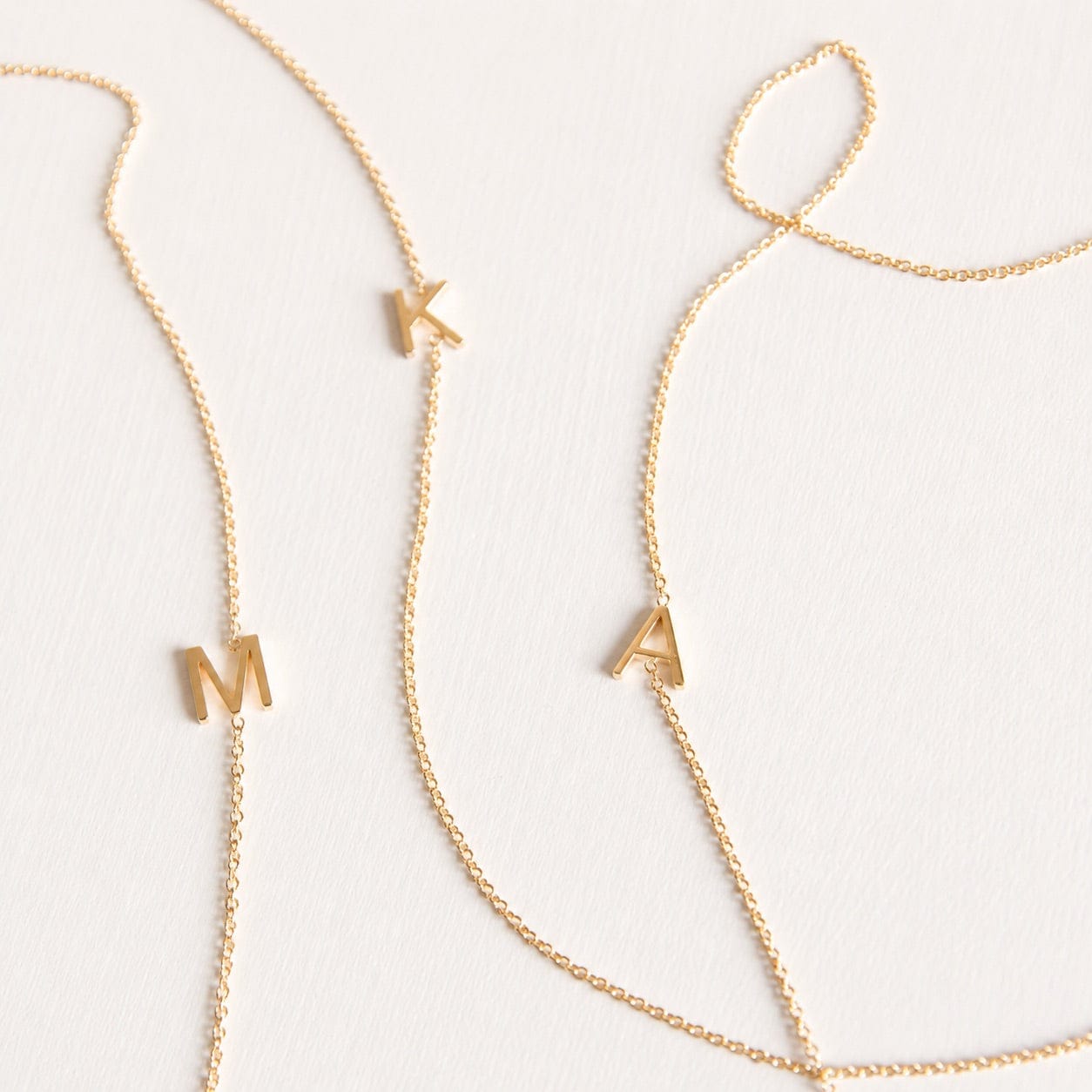 Three gold necklaces with single letter charms, being M, K and A on a plain background. 