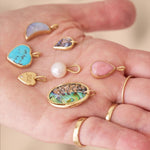 Hand holding various colorful gemstone pendants and a pearl on a neutral background
