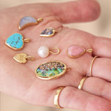 Hand holding various colorful gemstone pendants and a pearl on a neutral background
