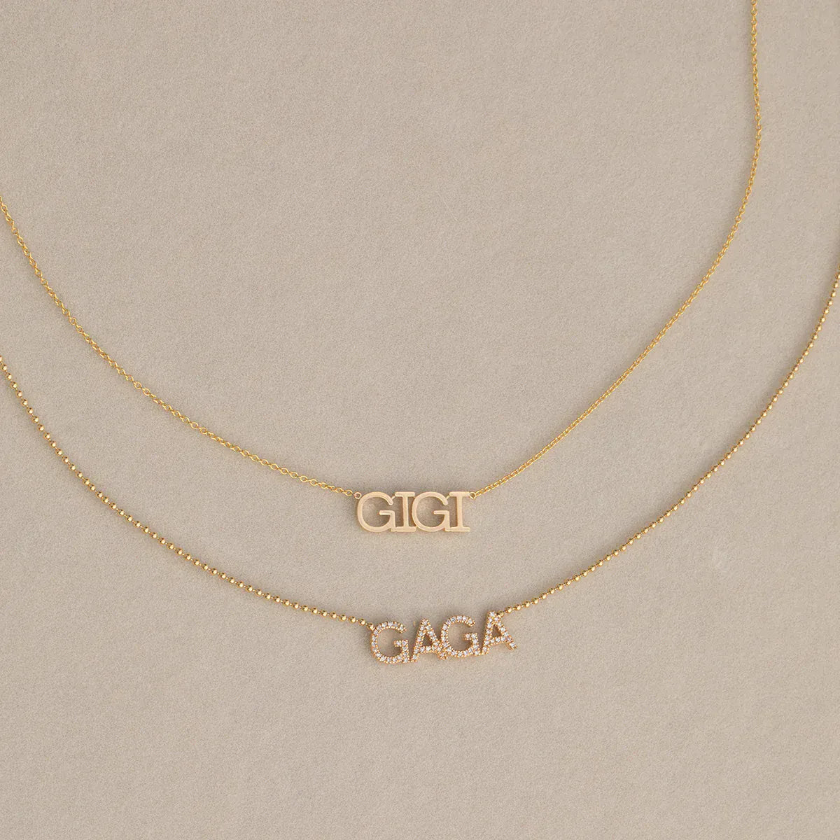 A GIGI Necklace in yellow gold with a cable chain and a Pavé Necklace in yellow gold with a bead chain, against a light beige background. 