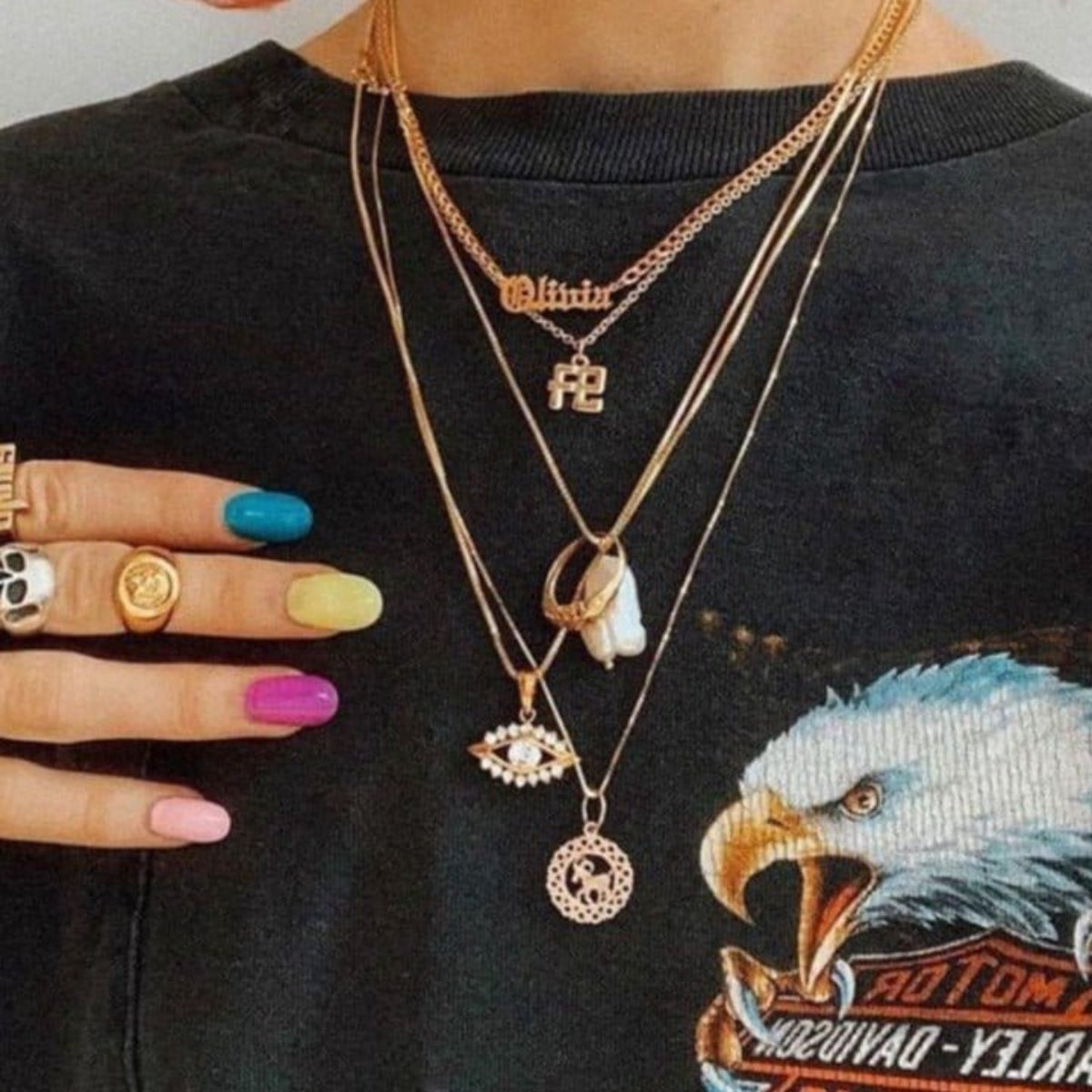 A person wearing multiple gold necklaces with various pendants, including a Zodiac Necklace - Aries, colourful nail polish, and a black shirt with an eagle design.