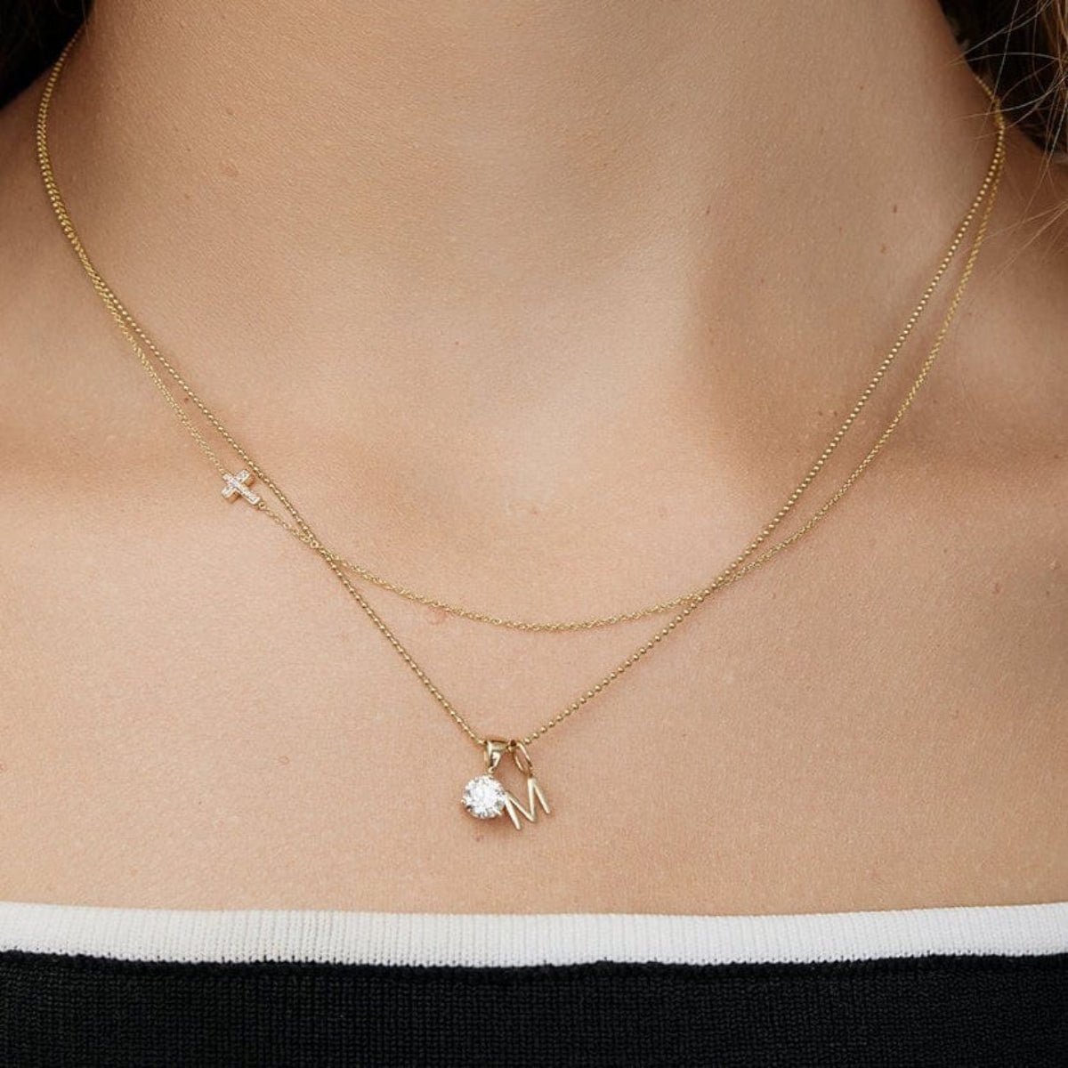 Close-up of a neck wearing a Classic Bead Chain with a Gold Letter Charm and Duchess Diamond Pendant, and a 14k Gold Asymmetrical Charm Necklace - Pave Cross, both in yellow gold. 