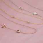 Three necklaces laid on a pink fabric background: a Monogram Necklace with Heart Rose Gold and a letter W charm, a Pavé Monogram Necklace with Diamond and a letter T charm and a Monogram Necklace with Heart White Gold and a letter S charm.