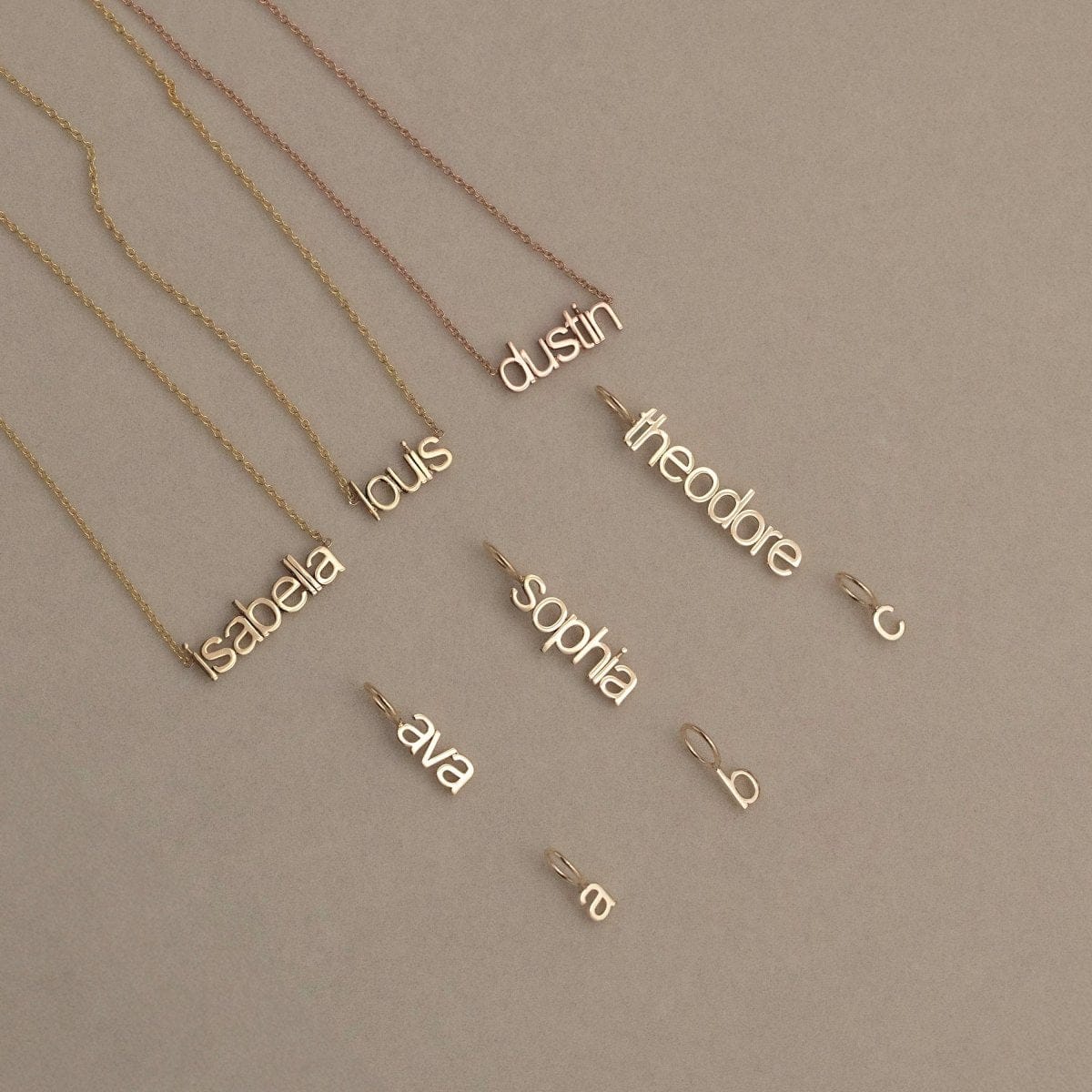 Three Petite Name Plate Necklaces with the names Isabella, Louis and Dustin, three Petite Nameplate Pendants in the names Ava, Sophia and Theodore, and three Petite Letter Charms in lowercase a, b and c, all in yellow gold,  against a beige background. 