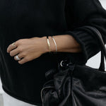 A close-up of an arm wearing a silver Flirt Bangle and a gold Plus One Cuff, a white gold Etched Paris Ring, an Open Diamond Ring and a C’est Moi Link Ring, styled with a black knit and holding a black bag.