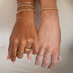 Two hands wearing several bracelets and rings in yellow and white gold: a Bold Link Bracelet, an Element Long Link Bracelet, two BFF Bracelets, a Down the Line Bracelet, a Love Set Match Bracelet, a Geo Stacking Ring, an Etched Paris Ring, a Link Ring and an Open Diamond Ring, against a neutral background.