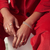 A pair of hands wearing a yellow gold Thick Band, Tres Pearl Ring,  Geo Stacking Ring Square, Delicate Beaded Band, Delicate Twist Band and two Tube Rings, along with mixed metal Rolo Bracelets and Diamond Cut Bracelets, styled in red corduroy clothing. 
