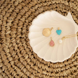 A Golden Scallop Shell, Etched Turquoise Heart Charm, Pink Opal Tear Drop Charm  and a Perla Necklace in a white shell holder against a textured woven surface. 