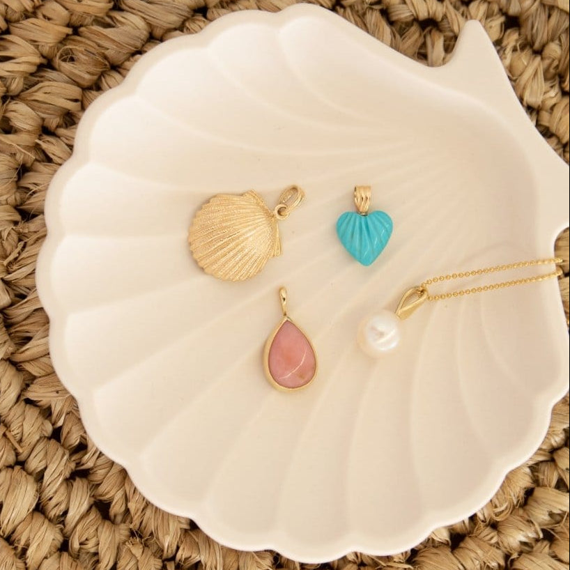 A Golden Scallop Shell, Etched Turquoise Heart Charm, Pink Opal Tear Drop Charm  and a Perla Necklace in a white shell holder against a textured woven surface. 
