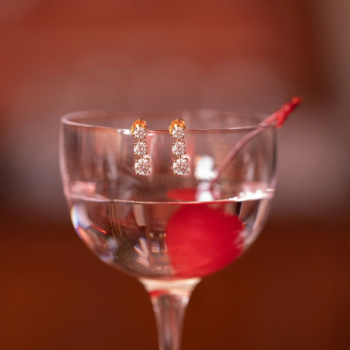 A pair of Triple Sec Diamond Earrings displayed in a cocktail glass with a red cherry against a blurred background. 