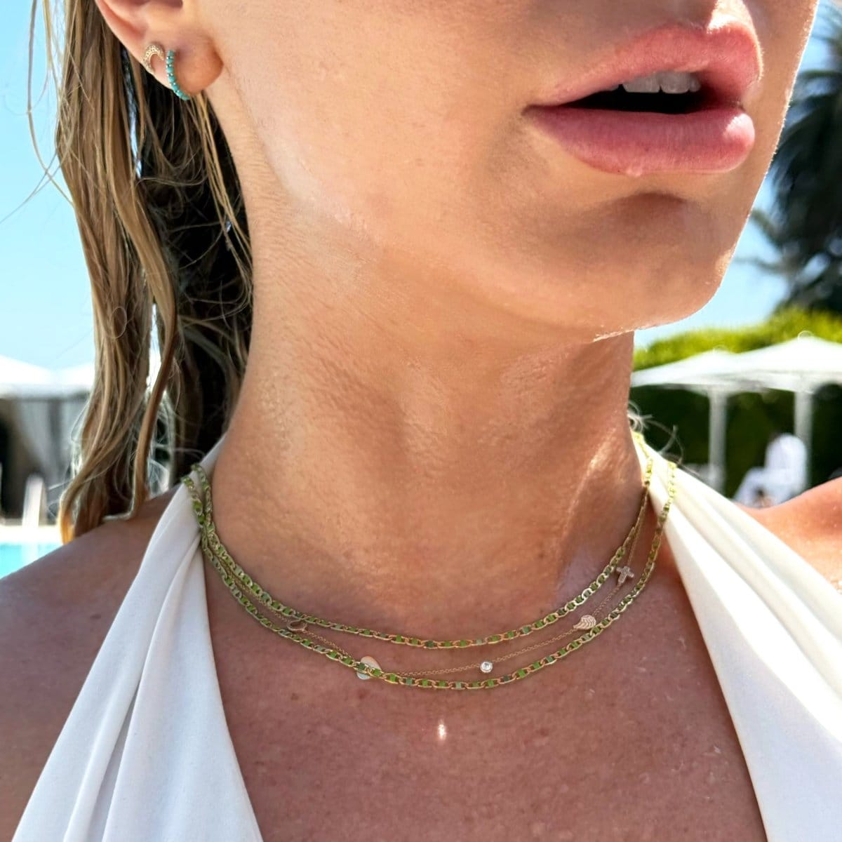 Close-up of a woman's neck wearing two Confetti Necklaces - Green, a Custom Gold Necklace - 5 Letters, an Alli Webb Turquoise Huggie and a Pavé Charm Stud - Moon, styled in a white halter top with blonde wet hair.