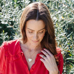 A brunette woman wearing a yellow gold Be Gentle in Grief Necklace along with several rings, including a Geo Stacking Ring Square, dressed in a red shirt against a green foliage background. 
