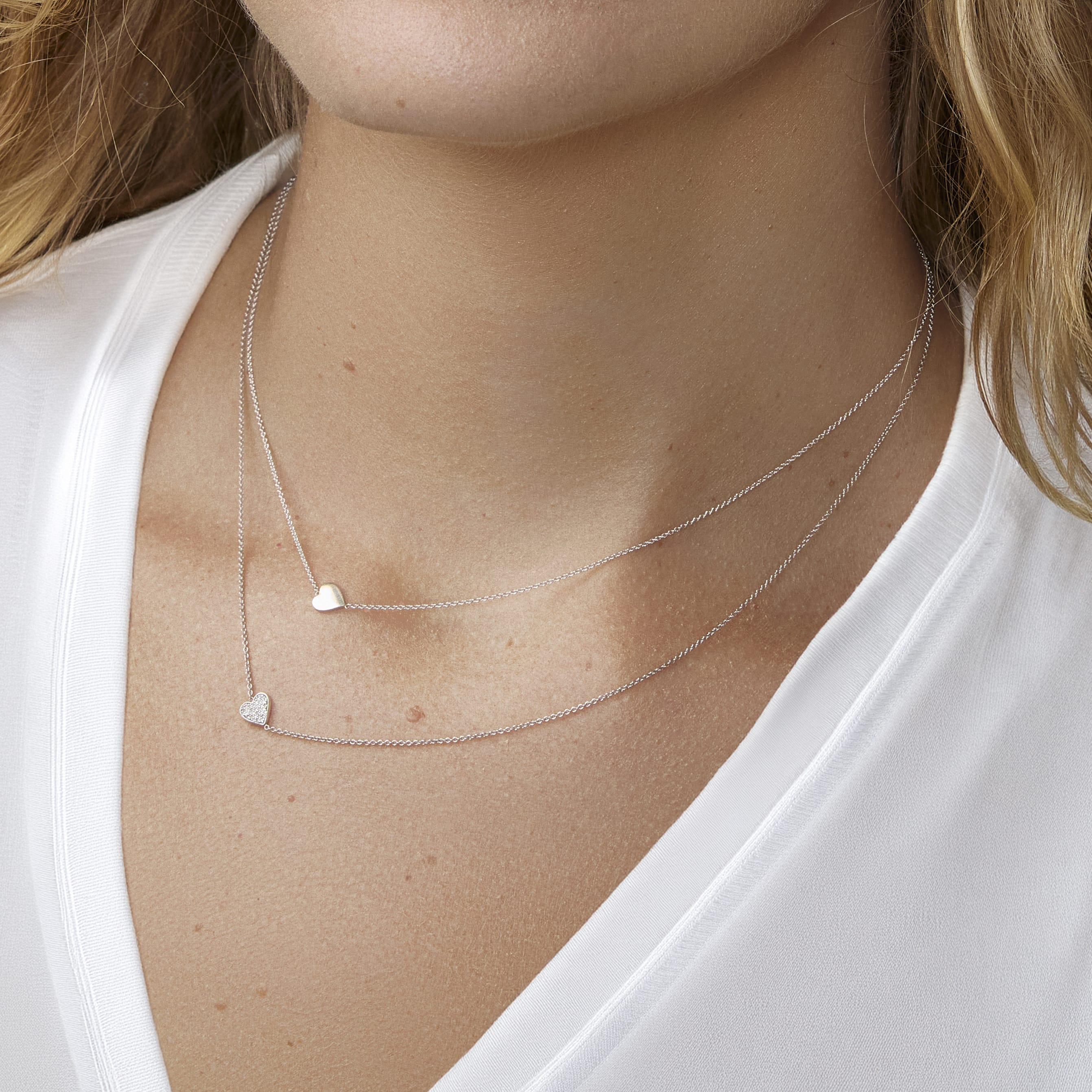 Close-up of a person wearing two silver necklaces with heart pendants on a white background