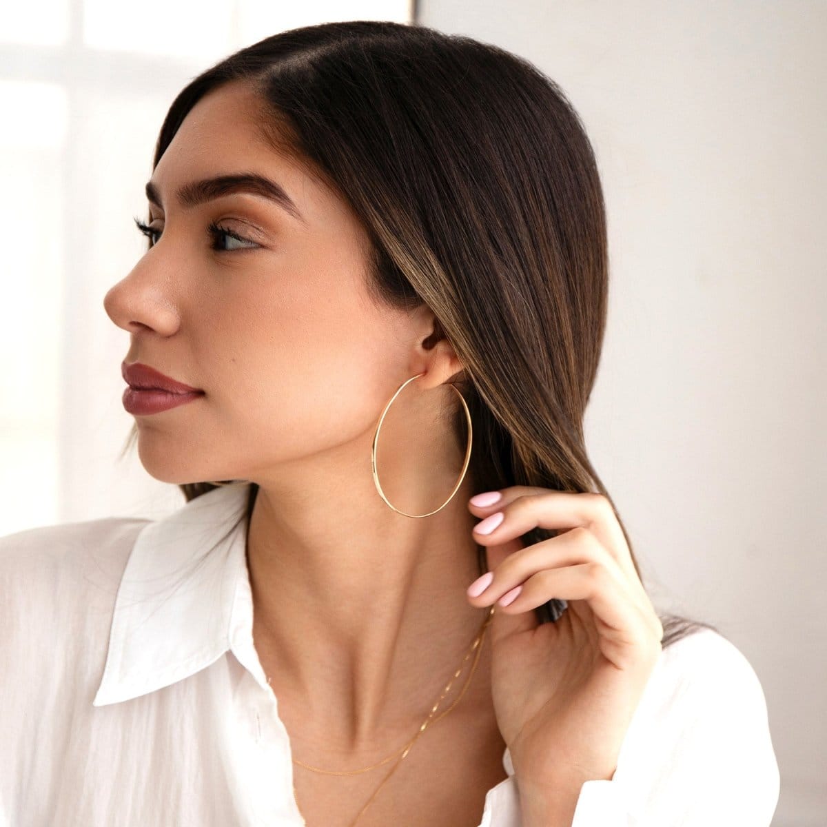 A woman wearing a Sleek & Chic Hoop - Large with two necklaces in yellow gold and a white shirt, with brown hair against a light background. 
