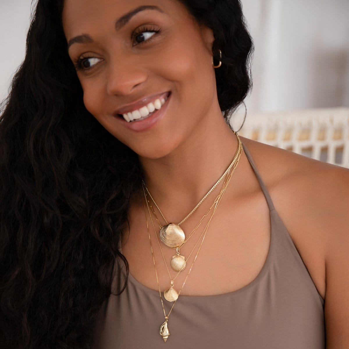 A woman wearing a Chic Cuban Curb Chain with The Golden Venus Shell, an Element Long Link Chain with a Golden Scallop Shell, a Classic Bead Chain with a Golden Coast Shell, The Slide Chain with a Golden Spiral Shell, with a light brown singlet top against a blurred background. 