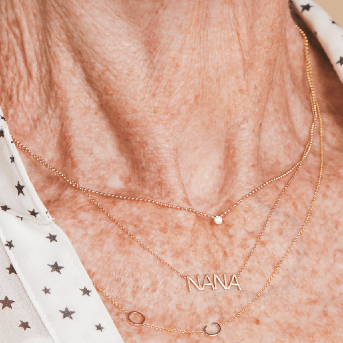 A close-up of a neck wearing a Single Diamond Layering Necklace, a NANA Necklace and a Custom Gold Necklace - 2 Letters, with a white top featuring black stars.