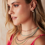 A blonde woman wearing The Peony Bouquet Necklace, The Bluebell Bouquet Necklace, The Thistle Bouquet Necklace, The Closer Bouquet Necklace and The Sage Bouquet Necklace, a Charmed I'm Sure Hoop / Heart and a Twisted Eclipse Stud, with a dark pink top against a neutral background. 