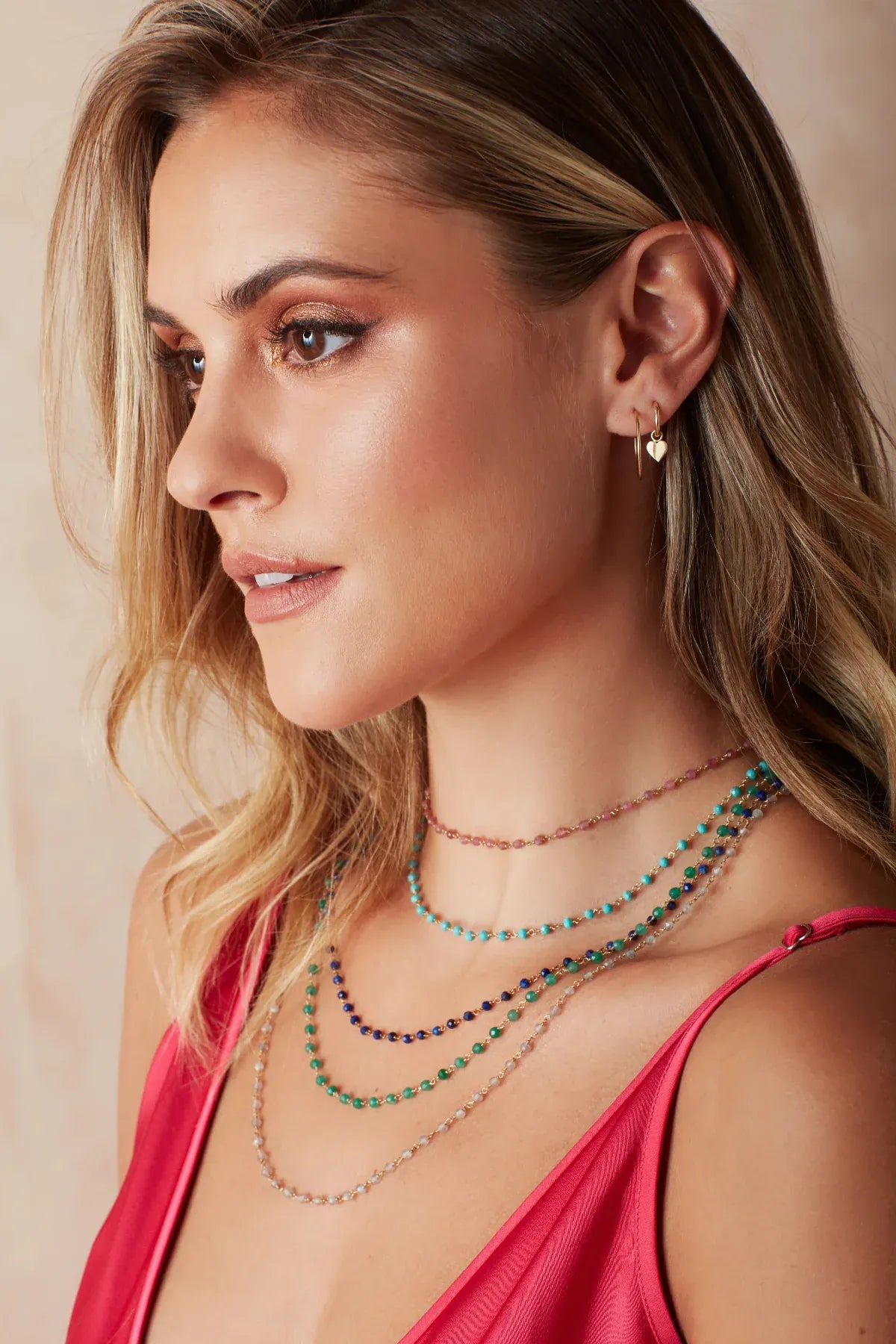 A blonde woman wearing The Peony Bouquet Necklace, The Bluebell Bouquet Necklace, The Thistle Bouquet Necklace, The Closer Bouquet Necklace and The Sage Bouquet Necklace, a Charmed I'm Sure Hoop / Heart and a Twisted Eclipse Stud, with a dark pink top against a neutral background. 