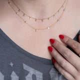 A close-up of a woman’s neck wearing a Stardust Diamond Necklace and a Custom Gold Necklace in yellow gold with seven charms (six birthstones and a pavé letter L) and a grey top, with her hand resting on her chest and red nails.