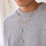 A close-up of a person wearing a pave MOMMY Necklace, an Element Long Link Chain with a Turquoise Heart Charm and a Perla Necklace, styled in a light grey jumper. 