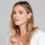 A woman wearing a Gold Safety Pin Earring, a Charmed I'm Sure Hoop / Star, two The Peony Bouquet Necklaces, The Sweetheart Necklace - Pink Sapphire, a Quintet Diamond Layering Necklace, and rings, all in yellow gold, with blonde hair and a white top against a light grey background.