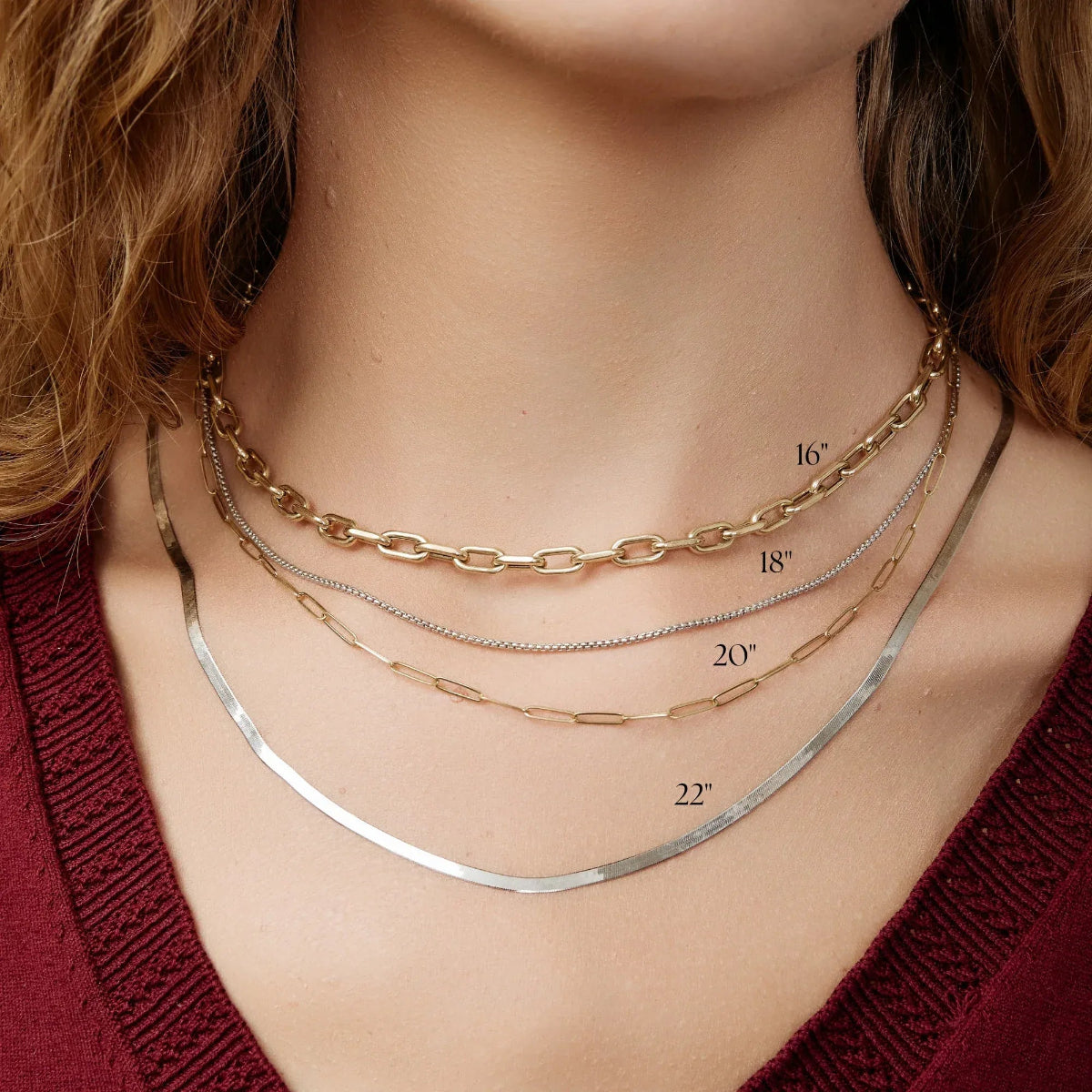 Close-up of a neck wearing a yellow gold Bold Link Chain, a white gold Box Chain, a yellow gold Element Long Link Chain and a white gold Sequence Chain, styled in a deep red top. 