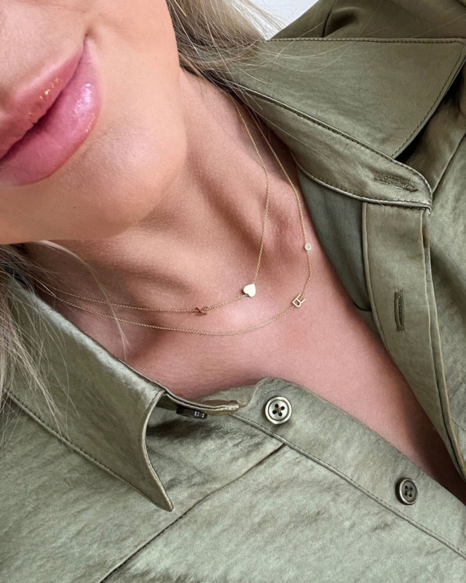 A close-up of a woman’s neck area wearing a Monogram Necklace with Heart with the letter K and a Monogram Necklace with Diamond with the letter R, both in yellow gold, in a sage green shirt.
