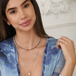 A brunette woman wearing Custom Gold Necklaces - 2 Letters in yellow gold, Endless Summer Necklaces in Coral Reef and Pink Sands, The Lily Bouquet Necklace, a Perla Necklace, a Perla Pinky Ring and a Thin Gold Hoop, with a blue top against white decorative background. 