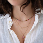 Close-up of a woman's neck wearing an Endless Summer Necklace - Coral Reef, Custom Gold Necklace - 5 Letters, a Classic Bead Chain with a Pink Opal Tear Drop Charm, styled in a white shirt. 