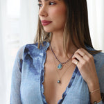A woman wearing the Thistle Bouquet Necklace with a Large Abalone Cabochon Charm, a Baroque Pearl Necklace, a Perla Pinky Ring, two Birthstone Rings, a Simple Bangle, and a Baroque Black Pearl Bangle, styled in a blue top against a white background.