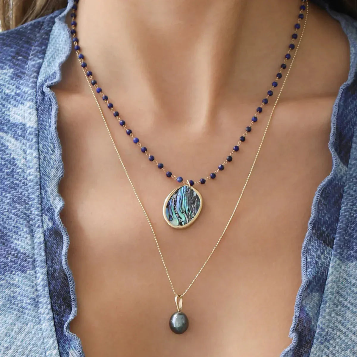 Close-up of a woman's décolletage wearing a Thistle Bouquet Necklace with a  Large Abalone Cabochon Charm and a Baroque Pearl Necklace, with blue top. 