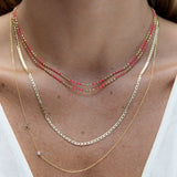 A close-up of a neck wearing two Confetti Necklaces - Pink, an Overboard Layering Chain and a Monogram Necklace with Diamond Yellow Gold and the letter K, styled in a white top. 