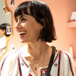 A smiling woman wearing layered necklaces, including the Courage Retreat Necklace, a FEMME Necklace, a Custom Gold Necklace - 2 Letters, and an Element Short Link Chain with an Abalone Leaf Charm, all in yellow gold, with short black hair and a multi-coloured top against a light pink background.