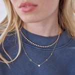 Close-up of a woman's neck wearing The Ultimate Diamond Necklace and a Custom Gold Necklace - 5 Letters, with a blue jumper and blonde hair. 