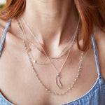 A close-up of a neck wearing an Element Long Link Chain, a Custom Gold Necklace - 2 Letters, a California Necklace, all in silver or white gold, and The Sage Bouquet Necklace, styled with a light blue denim top. 