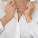 Close-up of a woman's decellotage wearing several The Lily Bouquet Necklaces with an Etched Turquoise Heart Charm, with her hands wearing The Lily Bouquet Bracelet, a Baroque White Pearl Bangle and a Perla Pinky Ring, in a white shirt. 