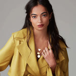 A brunette woman wearing a Geometric Circle Necklace layered with  similar necklaces featuring triangle and rectangle pendants, Sleek & Chic Hoops - Small, a Thick Band and a Tube Ring, all in yellow gold, with a mustard yellow jacket against a light grey background.