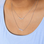 A close-up of a woman's décolletage wearing a Diamond Arc Layering Necklace and a MADRE Necklace, both in white gold, in a light blue top. 