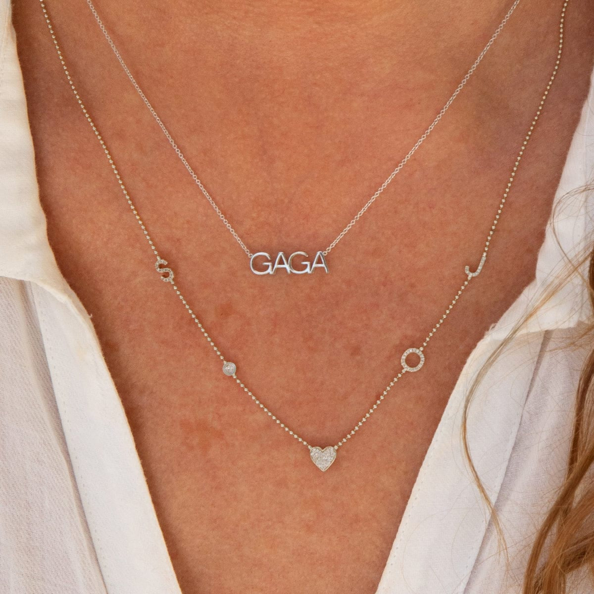 A close-up of a décolletage wearing a GAGA Necklace and a Custom Gold Necklace - 5 Letters, both in white gold and white shirt. 