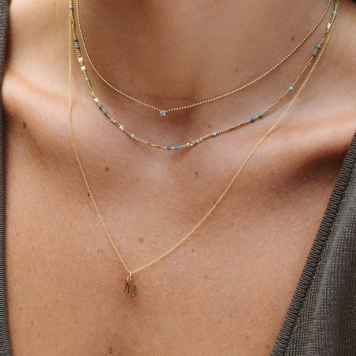 Close-up of a neck wearing a Single Diamond Layering Necklace, a Confetti Necklace - Blue and a chain necklace with a Gold Letter Charm in M, all in yellow or vermeil gold. 