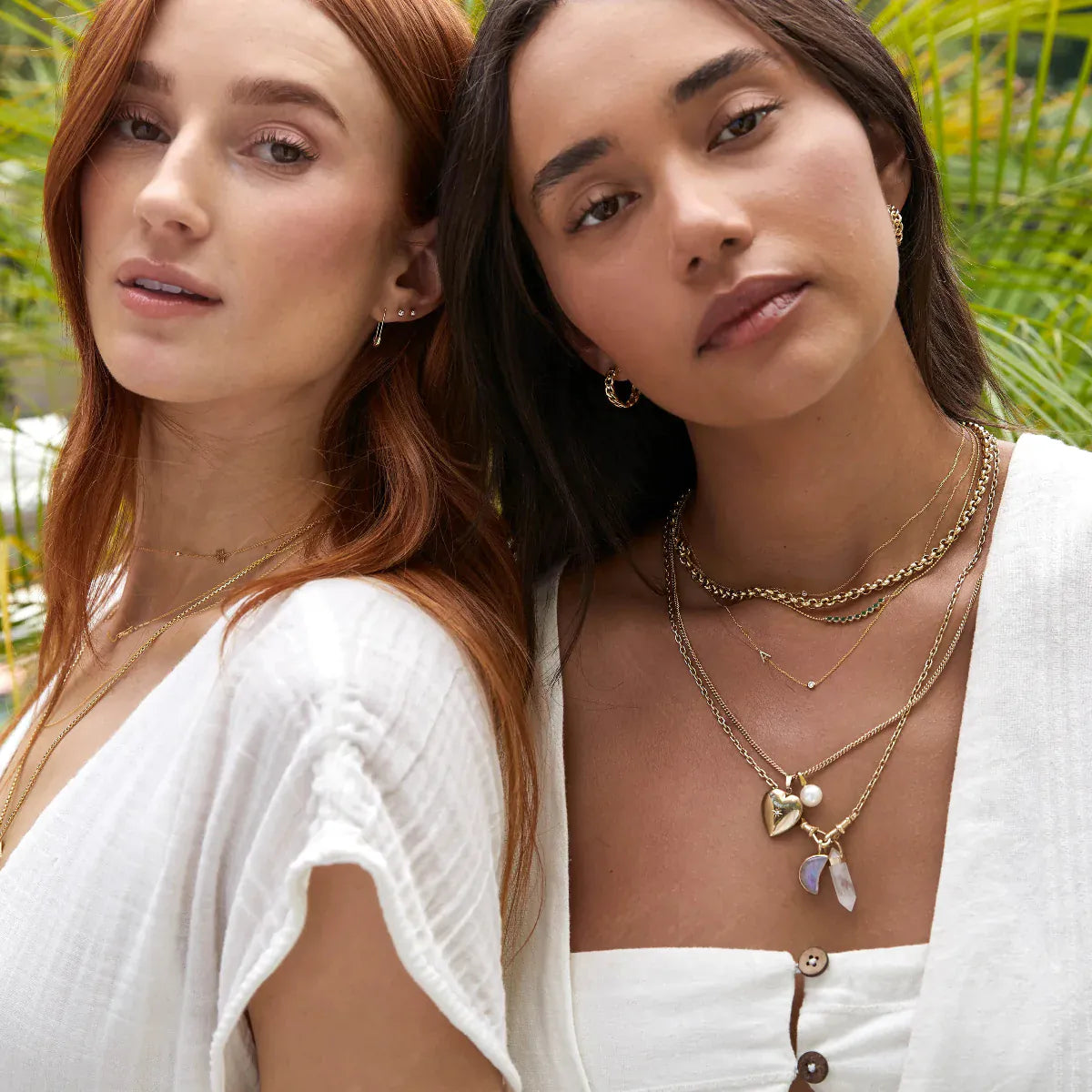 Two women, one wearing Link Hoops, a Rolo Necklace, Diamond Arc Layering Necklace, Monogram Necklace with Diamond, Box Chain with a Perla Pendant and Diamond Heart Locket Charm, Oval Link Chain with a Moonstone Crescent Charm and Balance Crystal Charm, and the other wearing a Gold Safety Pin Earring, two Birthstone Studs, Box Chain and other layered yellow gold necklaces, both wearing white tops against a leafy green outdoor background.