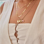 Close-up of a woman wearing a Custom Gold Necklace – 2 Letters, Overboard Layering Chain, Element Short Link Chain, Sequence Chain and Round Curb Chain with a Pink Opal Tear Drop Charm, Heart Locket Charm, Diamond Heart Locket Charm and Engraved Heart Charm, styled in a white linen top against a blurred outdoor background.