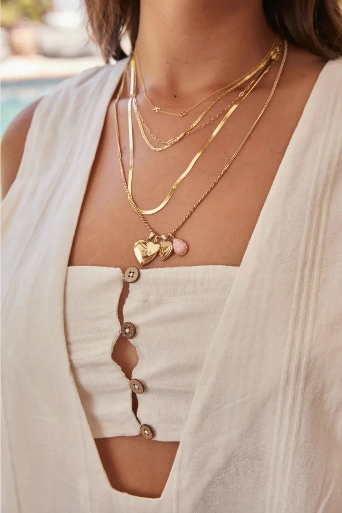 Close-up of a woman wearing a Custom Gold Necklace – 2 Letters, Overboard Layering Chain, Element Short Link Chain, Sequence Chain and Round Curb Chain with a Pink Opal Tear Drop Charm, Heart Locket Charm, Diamond Heart Locket Charm and Engraved Heart Charm, styled in a white linen top against a blurred outdoor background.